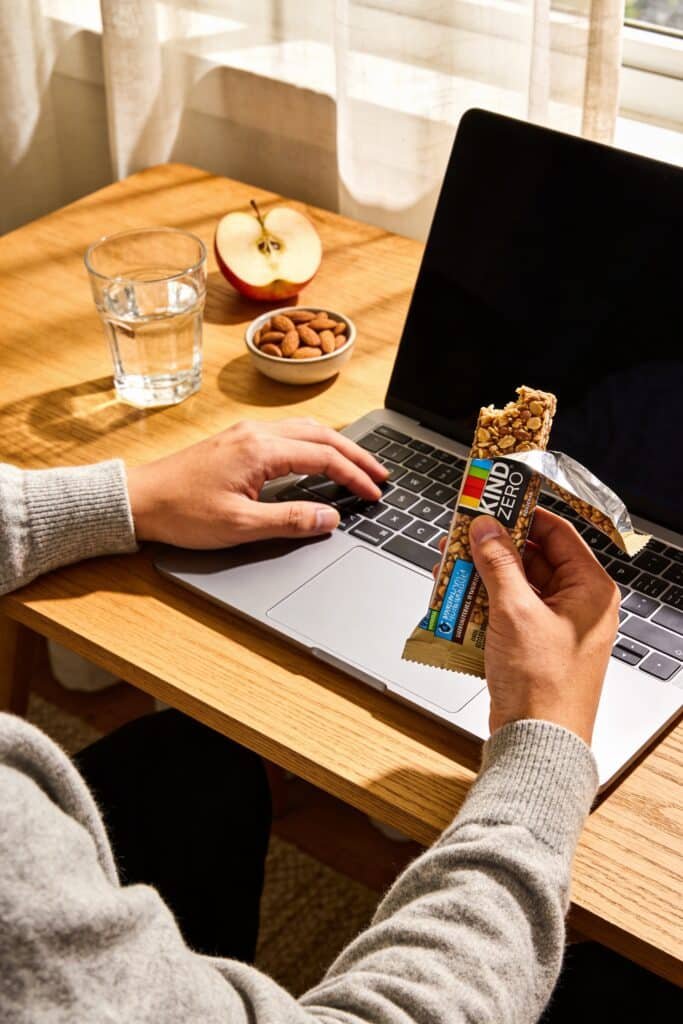 Lifestyle photography of a person enjoying a KIND ZERO bar while working at a laptop. Casual modern setting, healthy snacks on desk, morning light from window. Shows keto lifestyle integration. Warm, authentic stock photo style.