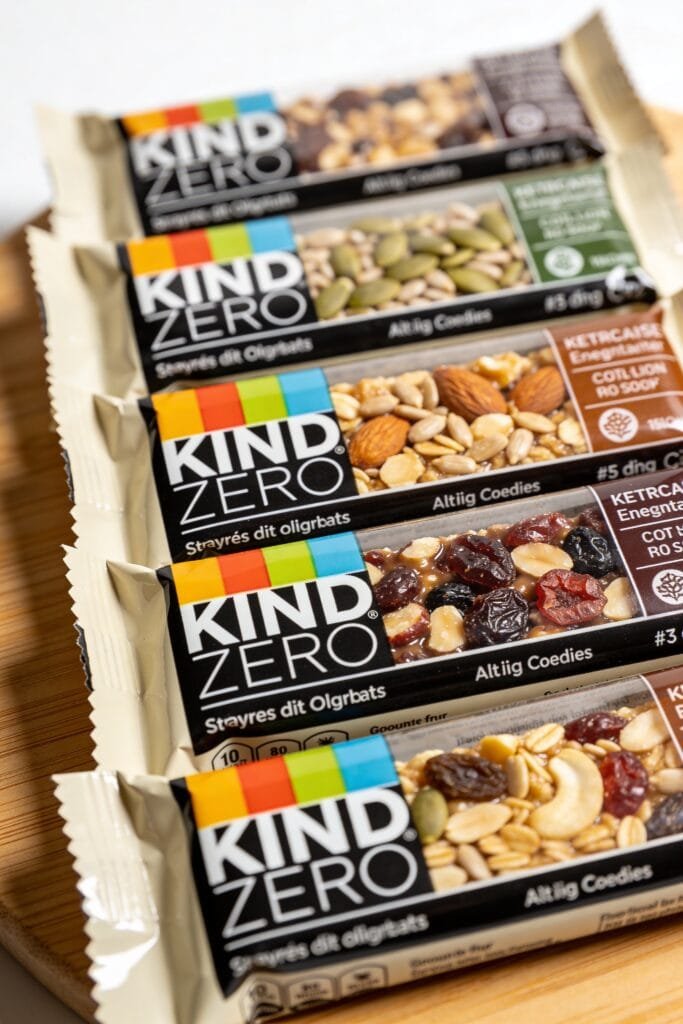 Professional product photography showing 5 KIND ZERO bar flavors side by side in a row. Each bar unwrapped to show interior texture. Labels visible. Clean white studio background, overhead lighting, food photography style.