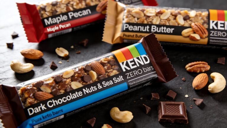 A flat lay photography composition of KIND ZERO keto bars arranged on a sleek dark surface. Dark Chocolate Nuts & Sea Salt bar in foreground with Maple Pecan and Peanut Butter flavors in background. Natural lighting from top-left, professional food photography style, shallow depth of field. Includes scattered nuts and chocolate pieces for texture.