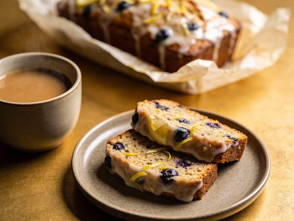 Two slices of glazed lemon blueberry keto bread served on a modern ceramic plate next to a cup of coffee. The slices are perfectly uniform. In the background, the rest of the loaf is wrapped in parchment paper. Cozy, inviting atmosphere, high-end lifestyle blog aesthetic.
