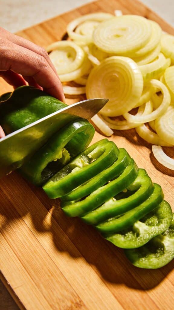 Subject: A close-up action shot of a chef's knife slicing through a vibrant green bell pepper on a wooden board.
Detail: Sliced yellow onions are piled to the side, cut significantly thinner than the peppers.
Focus: The uniform thickness of the pepper strips (approx 1/2 inch) to ensure even cooking.