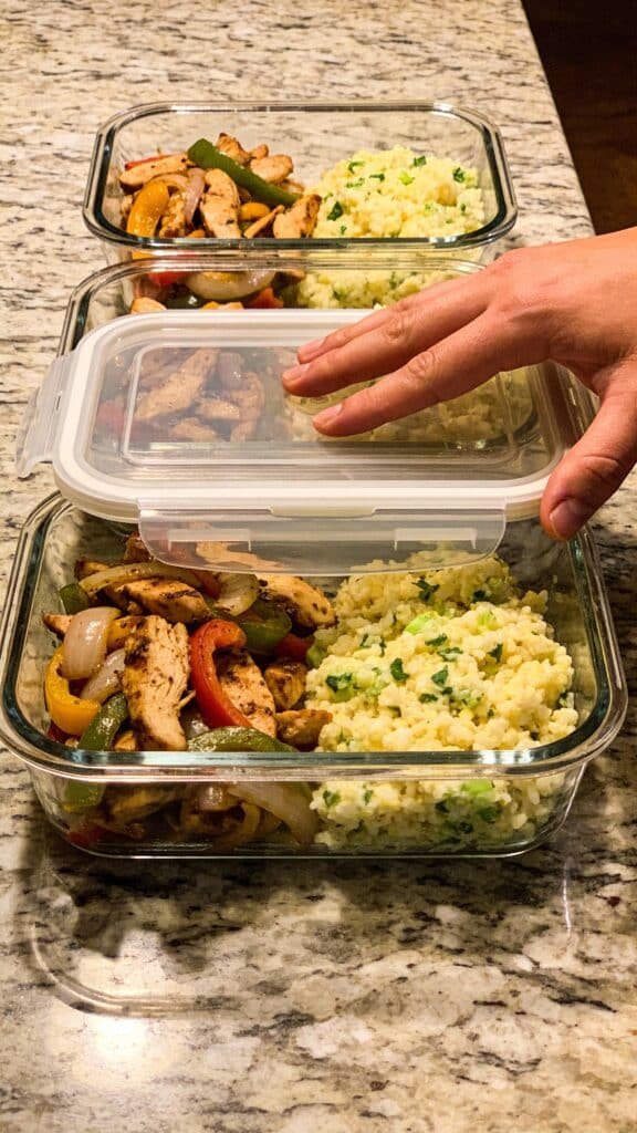 Subject: Three glass meal prep containers lined up on a granite counter.
Contents: Each container is filled with a portion of the cooked chicken fajita mixture and a side of fluffy cilantro-lime cauliflower rice.
Action: A hand is placing a snap-lock lid on the first container.