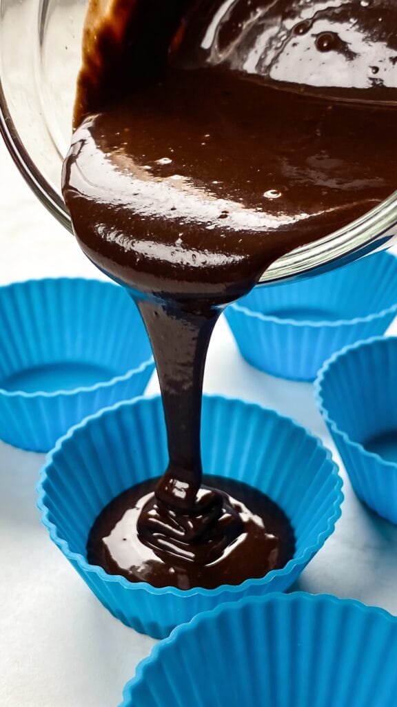 A close-up, overhead shot of dark, glossy melted chocolate being poured from a glass spout into bright blue silicone mini-muffin cups. The chocolate creates a smooth, mirror-like pool at the bottom.
